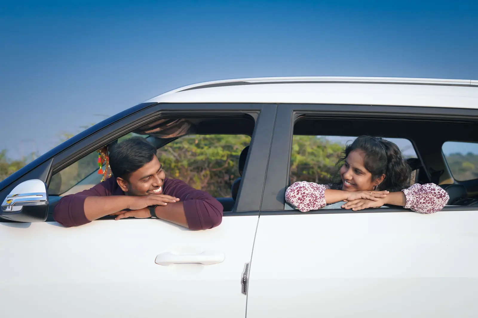 Two people smiling at each other from the front and back windows of a white car